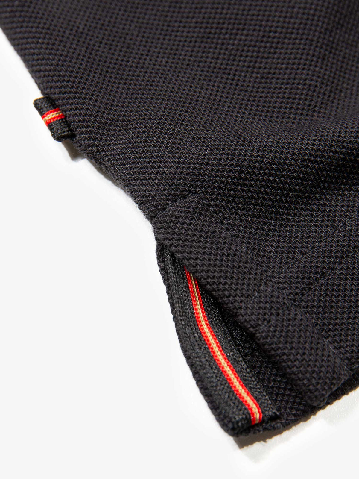 Close-up of side hem split with red trim on black New Willtape polo shirt