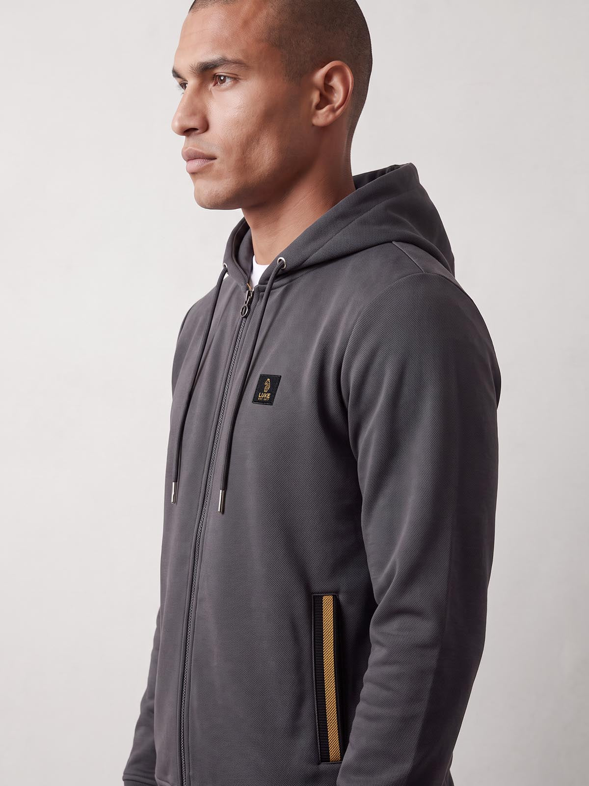 Close side view of man wearing charcoal LUKE 1977 Makati zip hoodie with woven badge and striped pocket