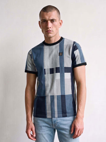 A man wearing a blue and gray checkered t-shirt with black trim, paired with light blue jeans, standing against a plain light gray background.